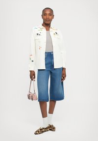 Woman wearing white embroidered jacket, gray top, knee-length denim shorts, leopard print slippers with white socks, and holding a small pink handbag.