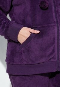Veste en polaire violette avec une surface texturée, fermeture éclair, poignets côtelés et une poche avant avec une main reposant à l'intérieur. Deux pompons sur la fermeture éclair.