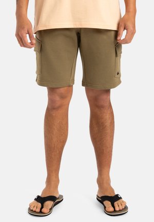 Hombre con shorts cargo verde oliva, camisa beige y chanclas negras, de pie con las manos relajadas a los lados sobre fondo blanco.