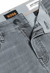 Grå denim jeans med en mjuk textur, med dragkedja, knäppning och kontrasterande sömmar. Etiketter visar "BOSS" och "SOFT MOTION."