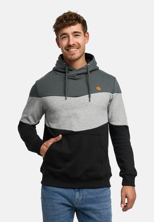 Młody mężczyzna uśmiechający się, ubrany w kolorową bluzę z kapturem w ciemnozielonym, szarym i czarnym kolorze, z jedną ręką w przedniej kieszeni, na jednolitym tle.