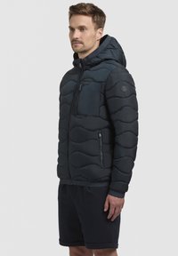 Navy pufferjack met capuchon, voorzien van een golvend quiltpatroon, ritszakken en elastische manchetten. Draag over donkere shorts.