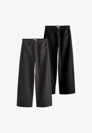 Person iført sorte jeans, grå top og beige jakke med den ene hånd i lommen, vist fra livet til midt på låret mod ensfarvet baggrund.