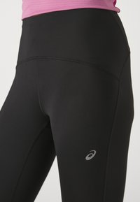 Juodos sportinės tamprės su aukštu liemeniu, lygiu audiniu ir sidabriniu logotipo akcentu. Turi plokščias siūles patogumui ir aptakų dizainą.