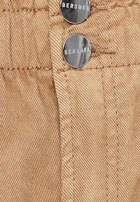 Tejido de color beige claro con canalé vertical, que presenta dos botones de metal plateado grabados con "BERSHKA" y detalles de costura visibles.