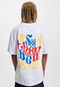 White cotton t-shirt featuring bold, multicolored text on the back: ��“See it then be it" in red, blue, and yellow with a sun graphic.