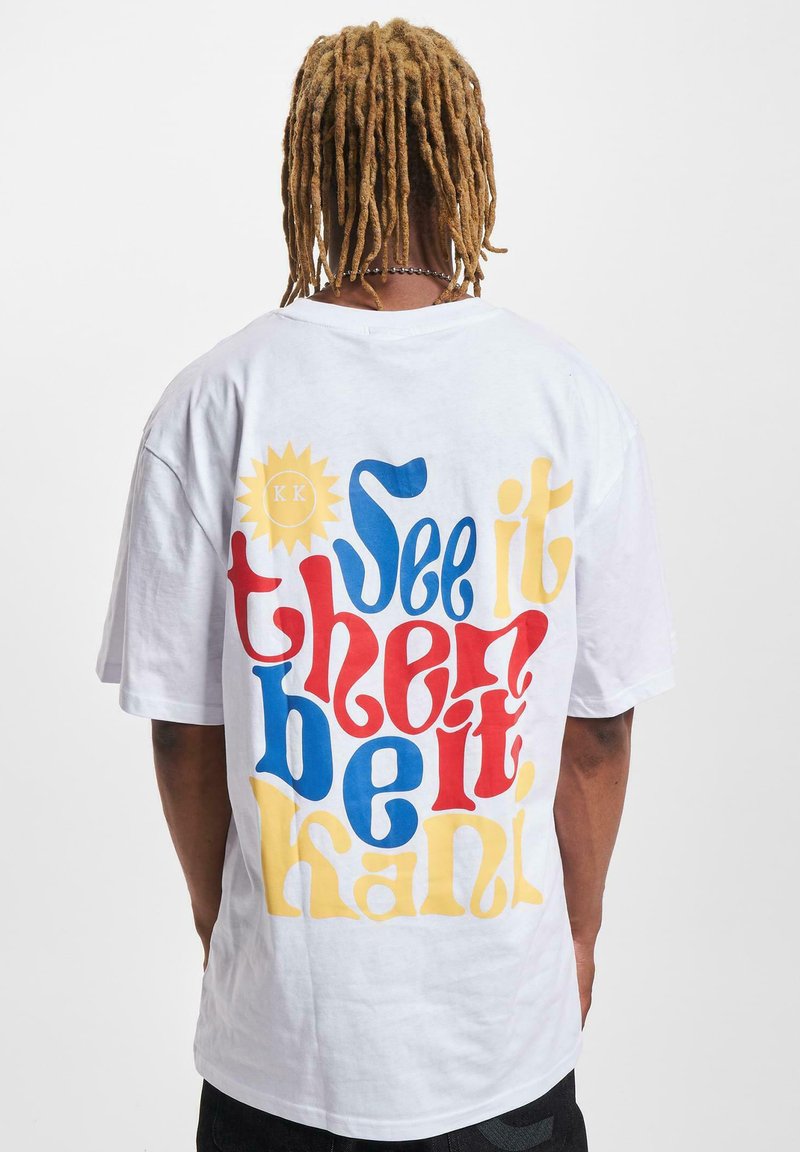 White cotton t-shirt featuring bold, multicolored text on the back: “See it then be it" in red, blue, and yellow with a sun graphic.
