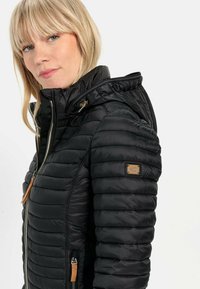 Sort puffervest med vandret quiltet tekstur, lynlås lukning og hætte. Har en kunstlæderpatch på ærmet og lynlåsdetaljer.