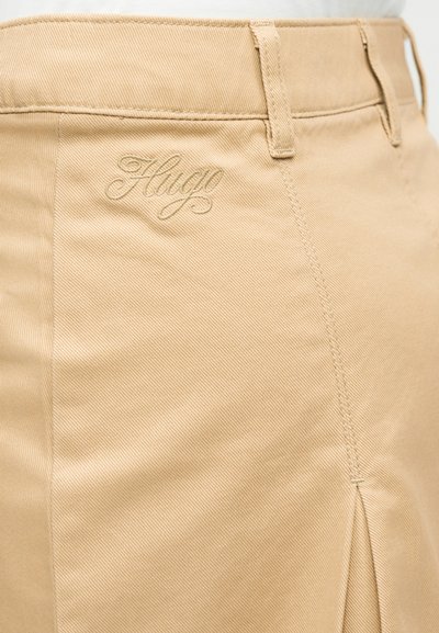 Gros plan sur un pantalon beige avec passants de ceinture et logo brodé "Hugo" près de la taille, montrant la texture du tissu et les détails des coutures.