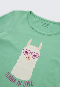 Grünes Kinderhemd mit einem weißen Lama, das rosa herzförmige Brillen trägt, und dem Text "LLAMA IN LOVE" in Pink darunter.