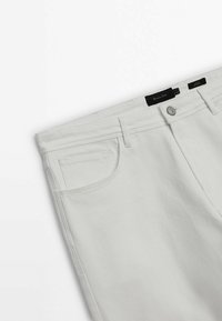 Pantalon en denim blanc avec poche avant, passants pour ceinture, bouton argenté et deux étiquettes en tissu noir indiquant la taille et la marque à la ceinture.