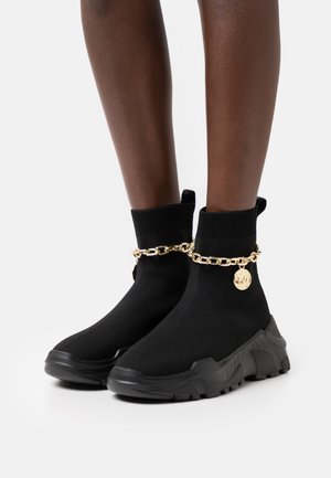 Sneakers nere in stile calza con suole spesse indossate su gambe, con catena dorata e medaglione rotondo come dettaglio intorno alle caviglie.