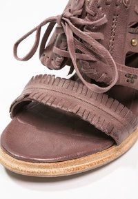 Gros plan d'une sandale en cuir mauve avec une bride à franges, un détail lacé, et une semelle texturée naturelle.