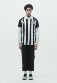 Svart og hvit stripete genser med lange ermer lagt over en hvit skjorte, sammen med løse svarte bukser og grå joggesko.