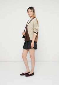 Junge Frau trägt einen beigen und braunen Cardigan, ein weißes Oberteil, einen schwarzen plissierten Rock und burgunderfarbene Ballerinas und steht vor einem schlichten weißen Hintergrund.
