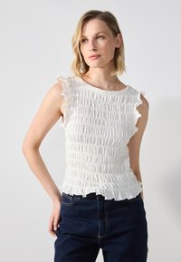 Hvit, ermeløs topp med smock-design, volanger langs skuldrene, og en tettsittende silhuett. Kombinert med mørkeblå jeans.