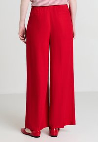 Weite rote Hosen mit elastischem Bund, zwei Gesäßtaschen und glatter Textur. Kombiniert mit roten flachen Schuhen.