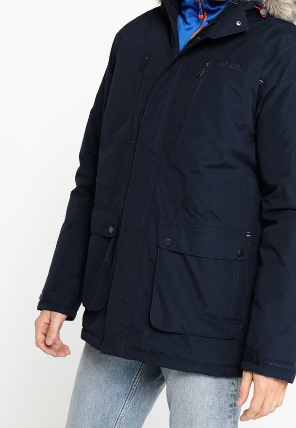 Homme portant une veste d'hiver bleu marine avec plusieurs poches zippées et boutonnées, un sweat à capuche bleu clair en dessous, et un jean délavé clair.