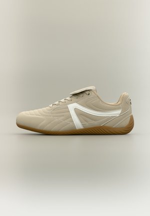 Beige sneaker with white side stripe, quilted texture, gum sole, and lace-up closure with a flap covering the laces.