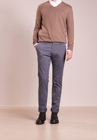 Pull en laine marron à col en V porté sur une chemise blanche à boutons, associé à un pantalon chino gris en coton et des bottes noires à lacets. Design uni, sans motifs.