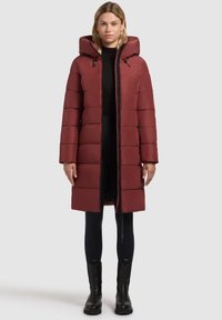Abrigo piumino rojo con capucha, diseño acolchado, largo, con cremallera frontal y bolsillos laterales; combinado con leggings negros y botines.