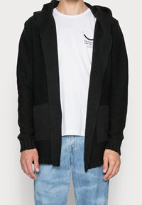 Svart stickad hoodie cardigan med ribbade manchetter och nederkant, med en framficka och öppen framsida, bärs över en vit T-shirt och blå jeans.