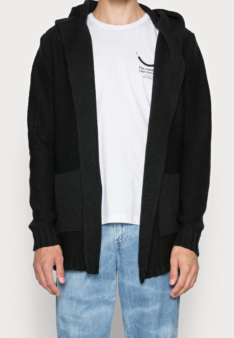 Svart stickad hoodie cardigan med ribbade manchetter och nederkant, med en framficka och öppen framsida, bärs över en vit T-shirt och blå jeans.