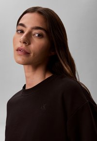Jeune femme aux longs cheveux bruns portant un sweat-shirt noir Calvin Klein, regardant légèrement par-dessus son épaule sur un fond gris uni.