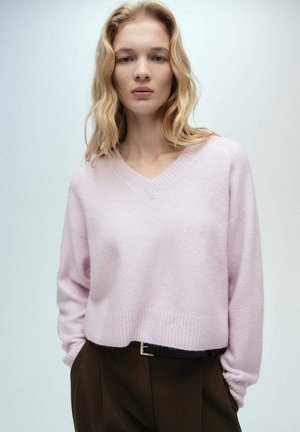 Lichtroze, cropped V-halstrui gemaakt van zacht materiaal, met ribbelstof aan de mouwen en onderkant, gecombineerd met bruine high-waisted broeken.
