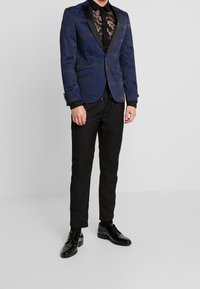 Giacca da smoking con motivo floreale blu e rever in satin nero, indossata sopra una camicia nera trasparente in pizzo e abbinata a pantaloni neri e scarpe lucide.