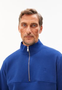 Blauwe fleece pullover met rits, gemaakt van zacht materiaal, met een hoge kraag, kenmerkend door een ritssluiting aan de voorkant en een klein geborduurd logo op de borst.