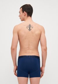 Boxers slip bleu marine avec une ceinture logo, texture lisse et design ajusté. Détail notable : un tatouage de fleur de lys.