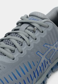 Γκρι αθλητικό παπούτσι με επάνω μέρος από δικτυωτό υλικό, μπλε λεπτομέρειες και υφή επιφάνειας. Διαθέτει κορδόνια, μαξιλαρωμένο κολάρο και διακόσμηση στο μεσαίο πέλμα με σχέδιο.