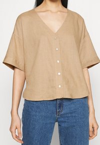 Camisa de lino beige con cuello en V, mangas caídas y cierre de botones en la parte delantera. Presenta una textura suave y un ajuste relajado.