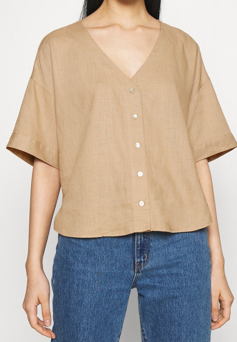 Camisa de lino beige con cuello en V, mangas caídas y cierre de botones en la parte delantera. Presenta una textura suave y un ajuste relajado.