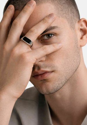 Uomo con capelli corti e barba corta, che copre parzialmente il volto con una mano che indossa un anello d'argento con una pietra nera, guardando attraverso le dita.