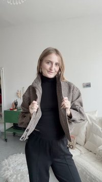 Brown and white houndstooth patterned jacket with a high collar, paired with a black turtleneck and loose black pants. Silver hoop earrings.