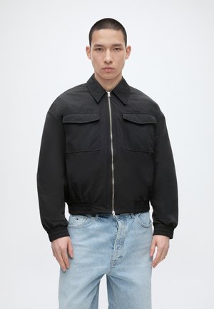 Férfi fekete cipzáras dzsekit visel elülső zsebekkel és világoskék farmerral, egy egyszínű világos háttér előtt áll.