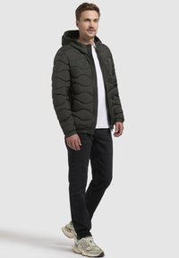 Dunkelgrüne gepolsterte Jacke mit Kapuze, die ein wellenförmiges gestepptes Design aufweist und einen Reißverschluss vorne hat. Kombiniert mit schwarzen Jeans und Turnschuhen.