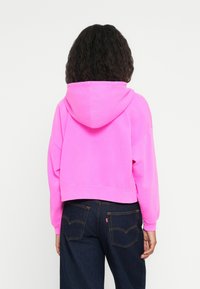 Felpa corta di un brillante colore rosa, realizzata in un tessuto morbido, presenta un cappuccio, maniche lunghe e polsini a coste; abbinata a jeans blu scuro.