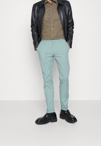 Mintgrüne maßgeschneiderte Hosen mit Seitentaschen, kombiniert mit einem braunen gemusterten Hemd und einer schwarzen Lederjacke. Schwarze Schuhe runden das Outfit ab.