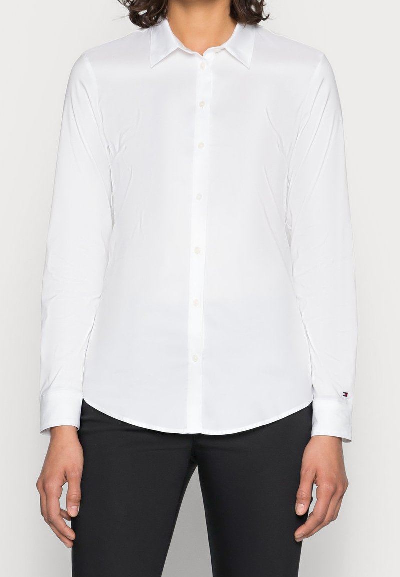 Personne portant une chemise blanche ajustée à manches longues boutonnée et un pantalon noir, debout devant un fond clair uni.