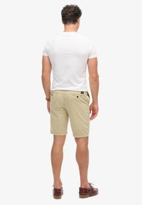 Pantaloni corti beige chiaro da uomo con una vestibilità dritta, dotati di tasche posteriori con dettagli rossi. Abbinati a una t-shirt bianca aderente.