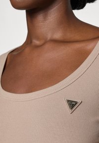 Béžový ribbed top s širokým výstřihem, který má trojúhelníkový kovový logo akcent u ramene. Hladký materiál, přiléhavý střih.