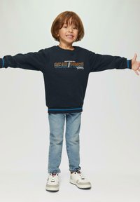 Felpa navy con grafica "GEAR//POWER", dettagli blu, abbinata a jeans azzurri e sneakers. Il bambino sorride con le braccia aperte.