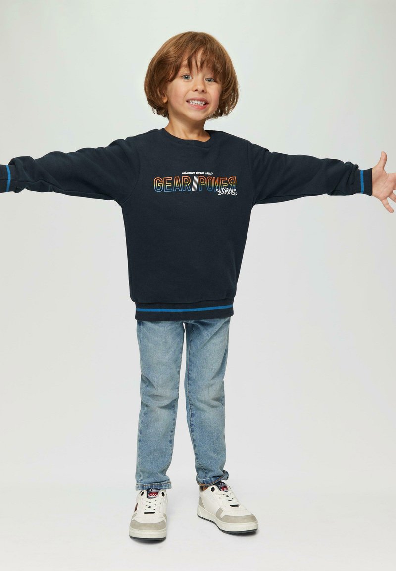 Felpa navy con grafica "GEAR//POWER", dettagli blu, abbinata a jeans azzurri e sneakers. Il bambino sorride con le braccia aperte.