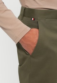 Zielone bawełniane spodnie z kieszeniami bocznymi i paskiem w talii z małą naszywką w czerwono-białe-niebieskie kolory. Widoczna jasnobrązowa koszula z długim rękawem.