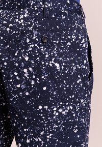 Niebieskie spodnie z tkaniny z plamami w białym i jasnoniebieskim wzorze. Posiadają tylną kieszeń z czarnym guzikiem i gładką fakturę.