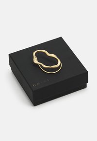 Goldene abstrakte Ring liegt auf einer strukturierten schwarzen Box mit goldener Markenkennzeichnung. Der Ring weist wellenartige, organische Formen und ein glänzendes Finish auf.