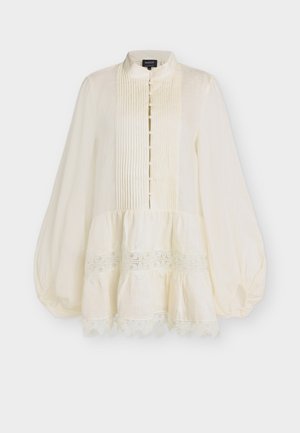 Μπεζ μπλούζα με μακριά μανίκια, πλισέ μπροστινό μέρος, κουμπιά καλυμμένα με ύφασμα, δαντελένιο τελείωμα, σούρες στο κάτω μέρος και φουσκωτά μανίκια.